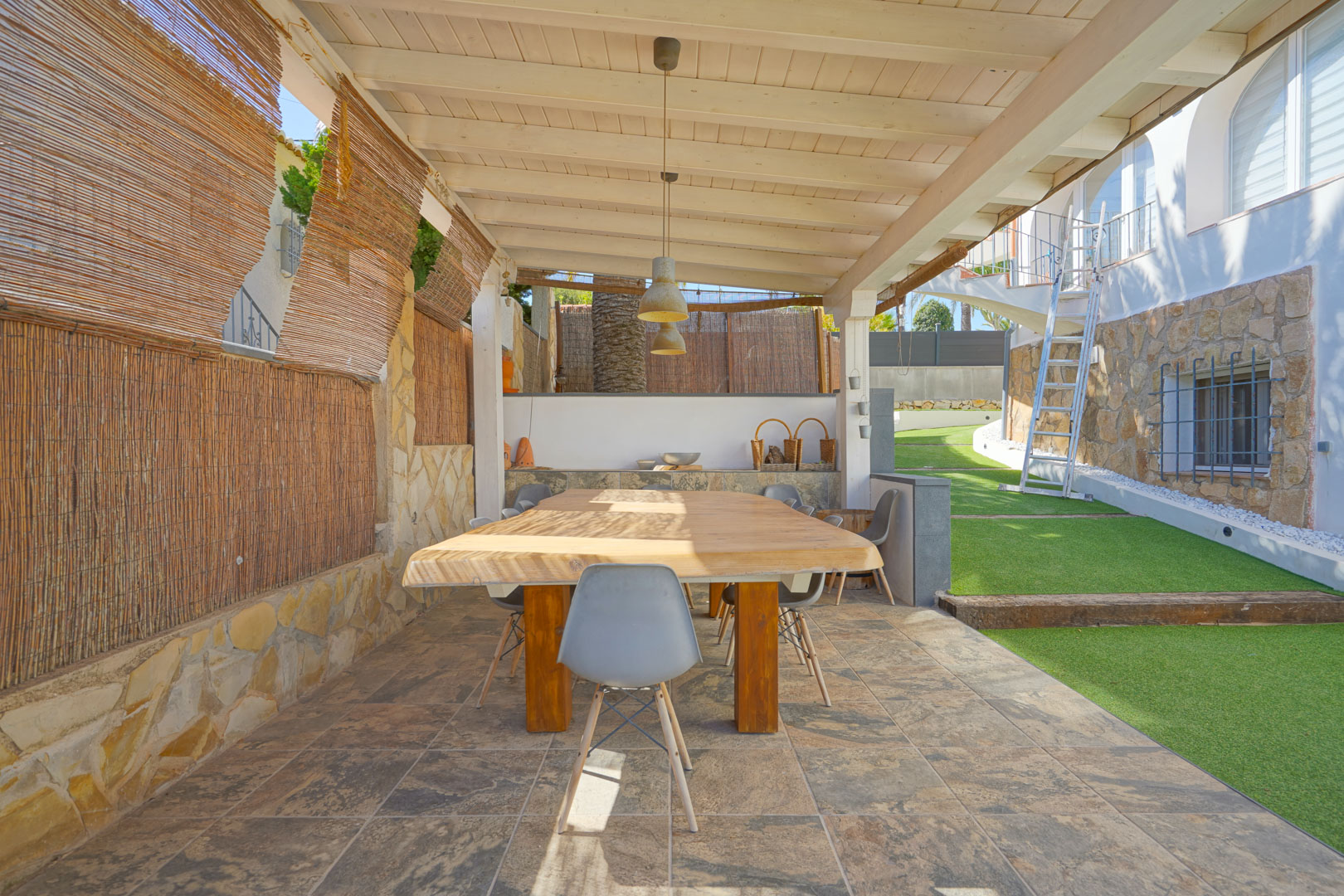 Outdoor dining area under a white wooden pergola, featuring a solid wood table, tiled flooring, and privacy screens, adjacent to a manicured lawn.