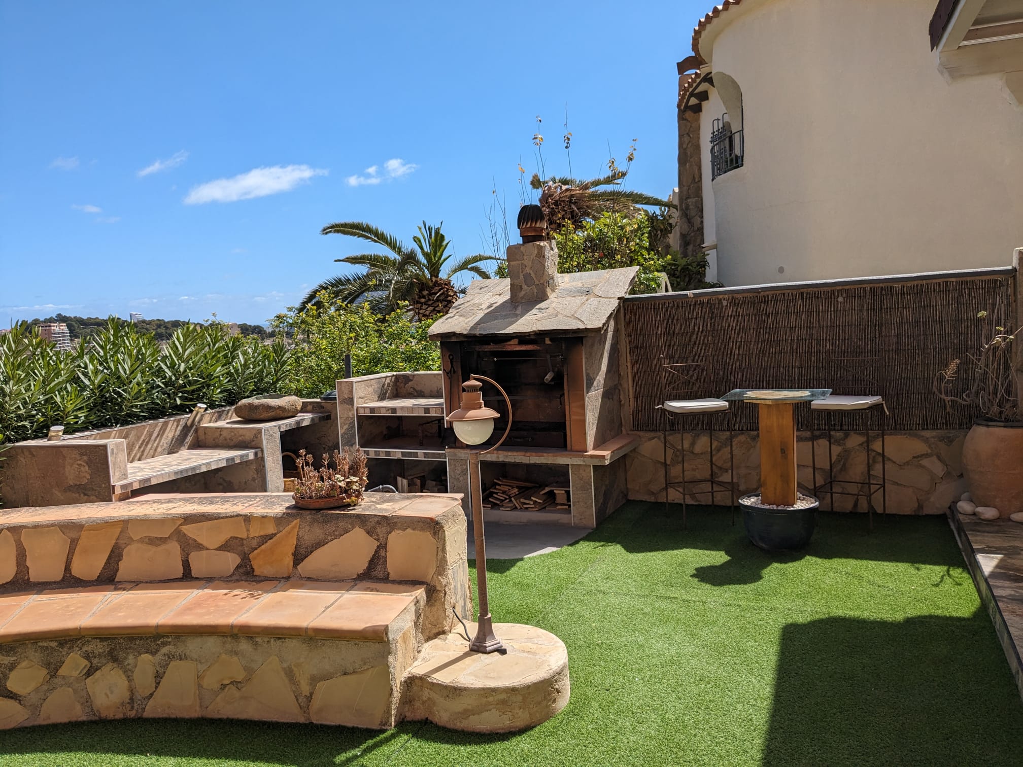 Outdoor stone barbecue area featuring artificial turf, built-in stone seating, and a high-top bar table with views of the Mediterranean coastline.