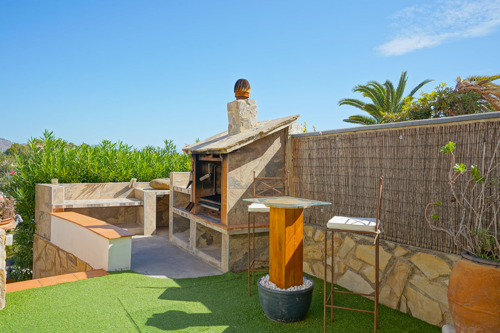 Outdoor stone-built barbecue area featuring tiled worktops, a wooden high-top table with glass, and bar stools set on an artificial lawn.
