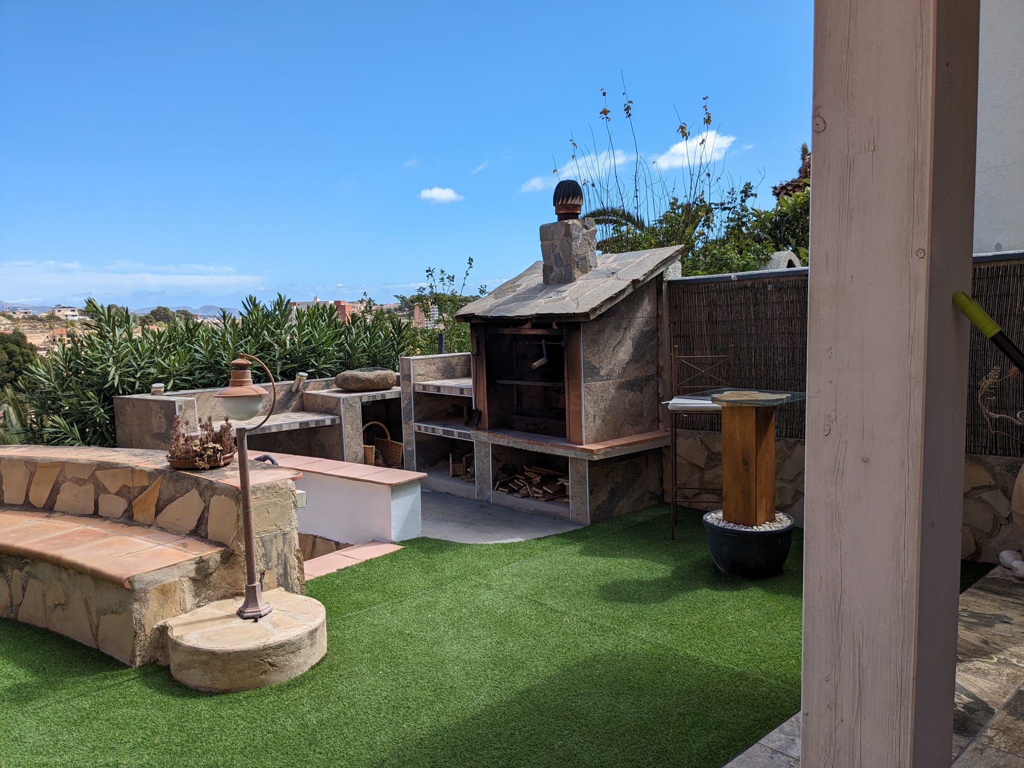 Outdoor stone-clad barbecue area featuring integrated shelving, a built-in stone bench with terracotta seating, and a clean artificial lawn.