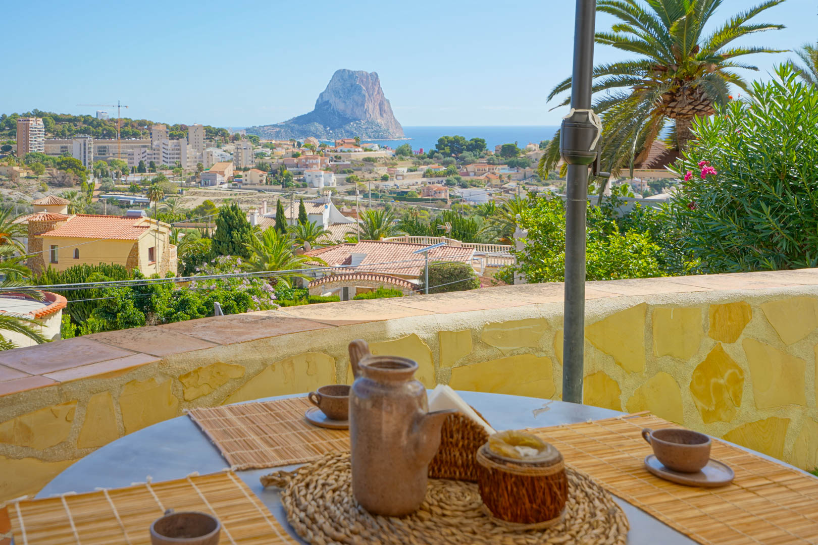 Outdoor terrace featuring a dining setup with panoramic views of the Peñón de Ifach and the Mediterranean Sea, framed by natural stone walls and palms.