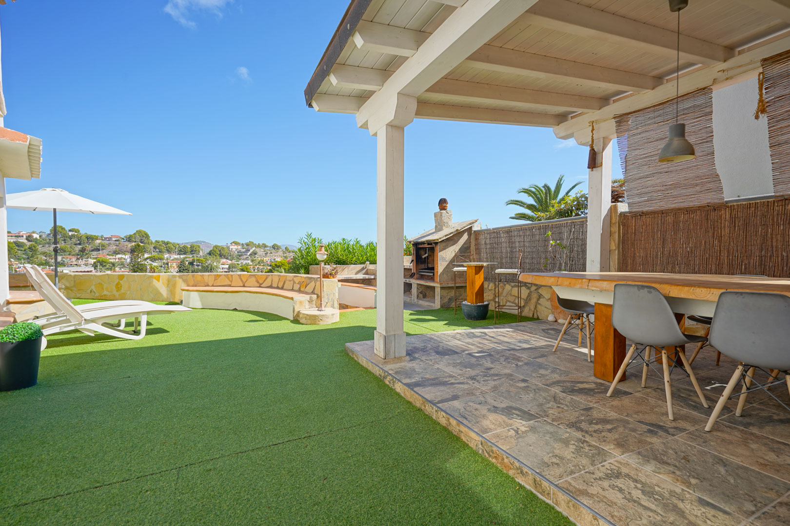 Outdoor terrace featuring a white wooden pergola, dining area, stone barbecue, and artificial turf section with sun loungers and panoramic views.