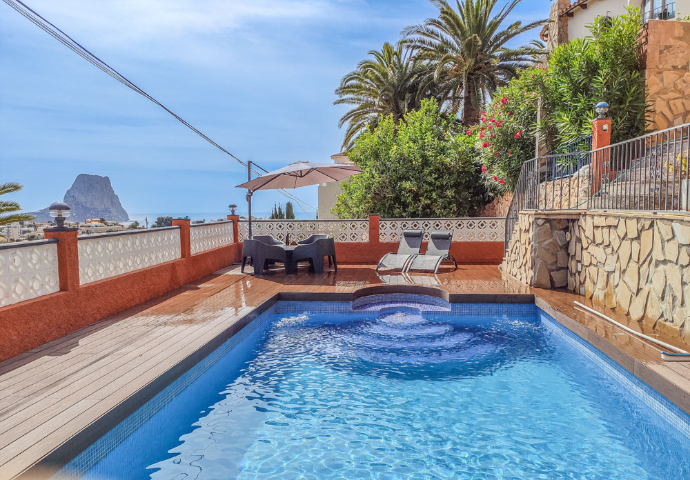 Outdoor terrace with a blue mosaic pool, wood-effect decking, and clear views of the Peñón de Ifach. Features a dining area with an umbrella and loungers.