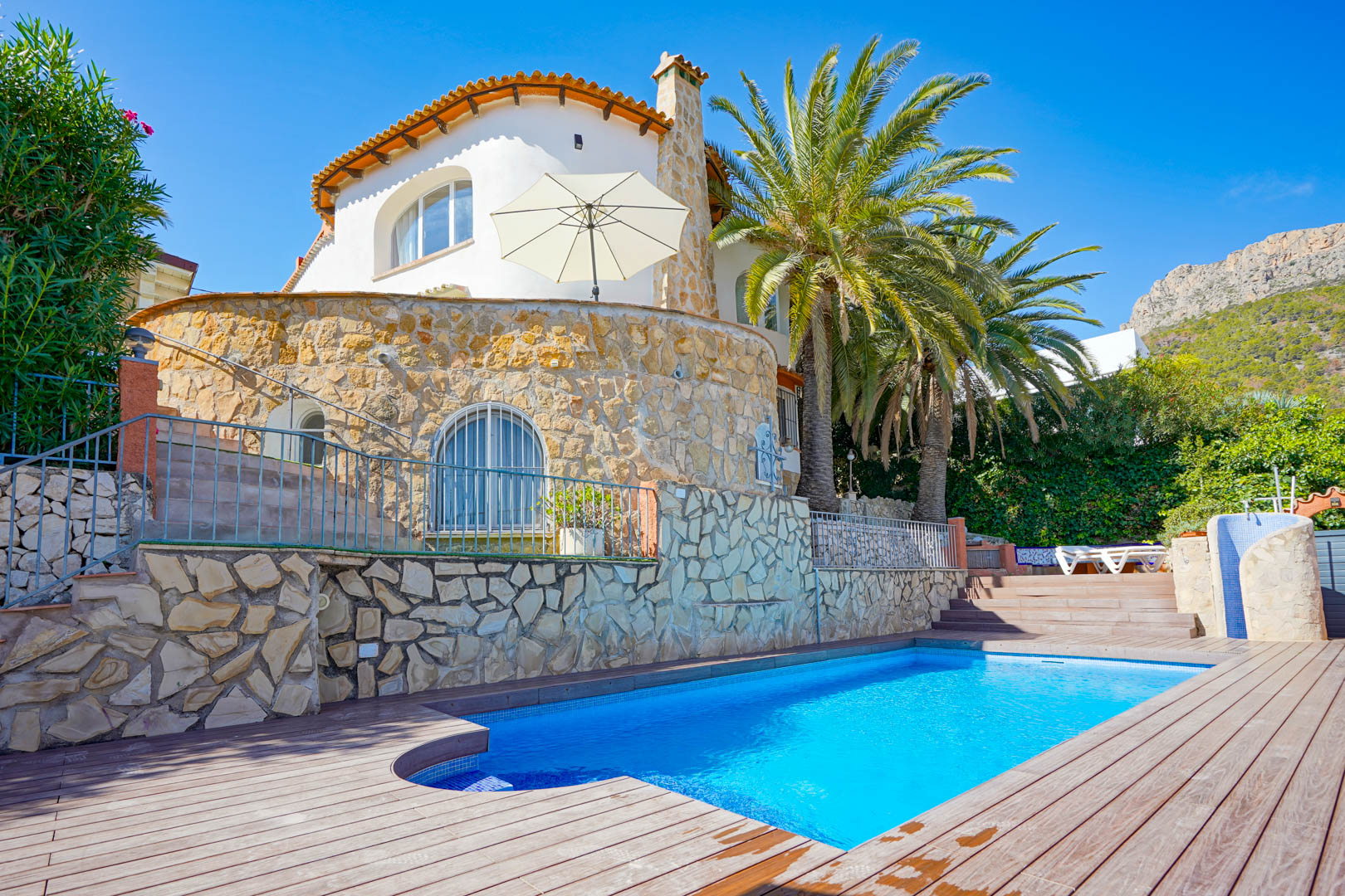 Private swimming pool featuring wooden decking and natural stone walls, complemented by mature palm trees and mountain views.