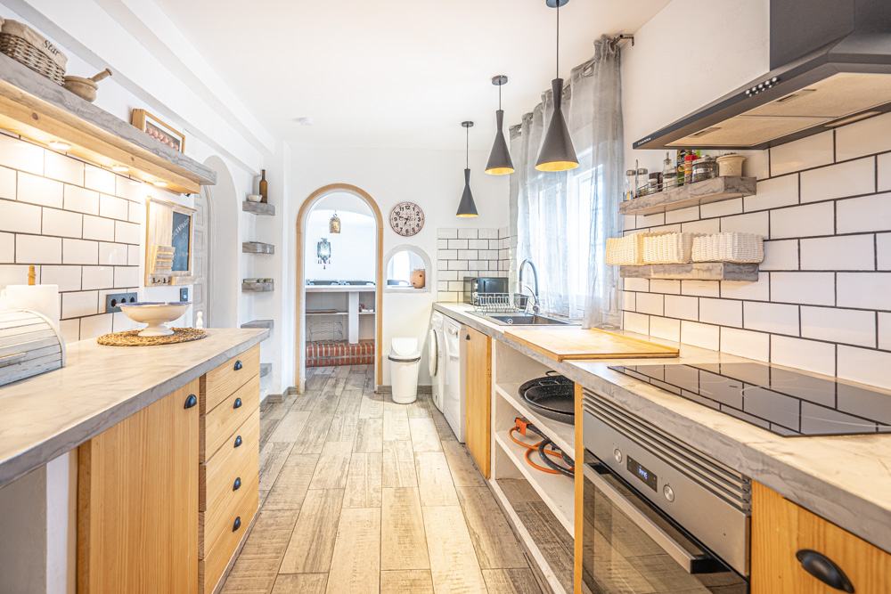 Rustic kitchen featuring grey countertops, wooden cabinetry, and white subway tiles. It includes integrated appliances, black pendant lighting, and an arched doorway leading to the dining area.
