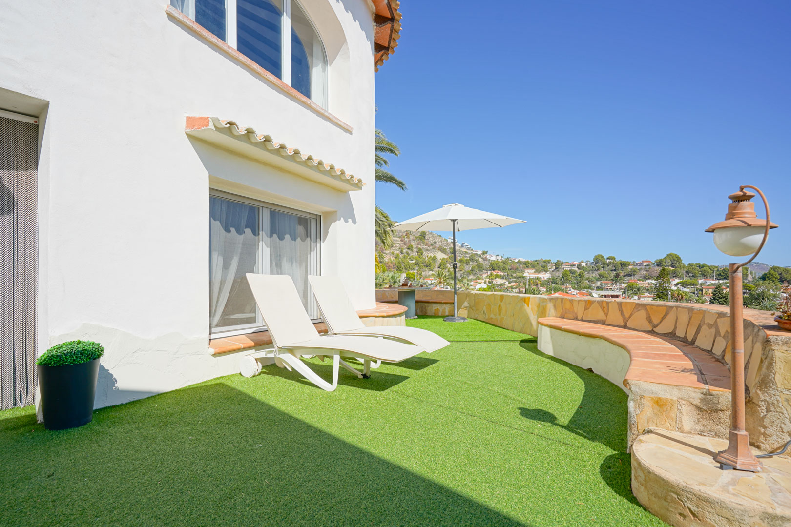 Terrace featuring artificial turf, white sun loungers, and a curved stone-clad perimeter wall with built-in seating overlooking the Calpe landscape.