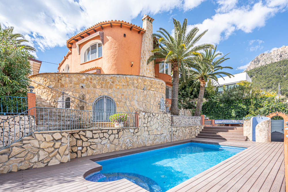 Villa exterior featuring a private pool with wood-effect decking, natural stone masonry, and a scenic backdrop of palm trees and mountains.