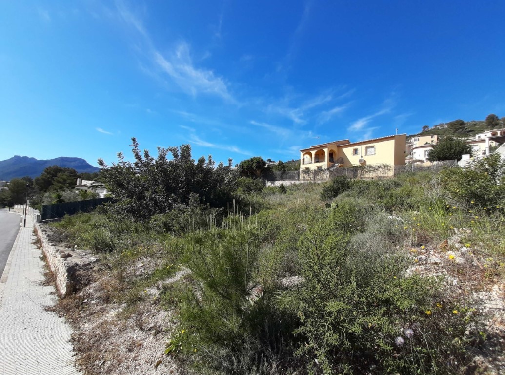 Plot featuring native Mediterranean vegetation and mountain views, located alongside a paved sidewalk in a quiet residential area.