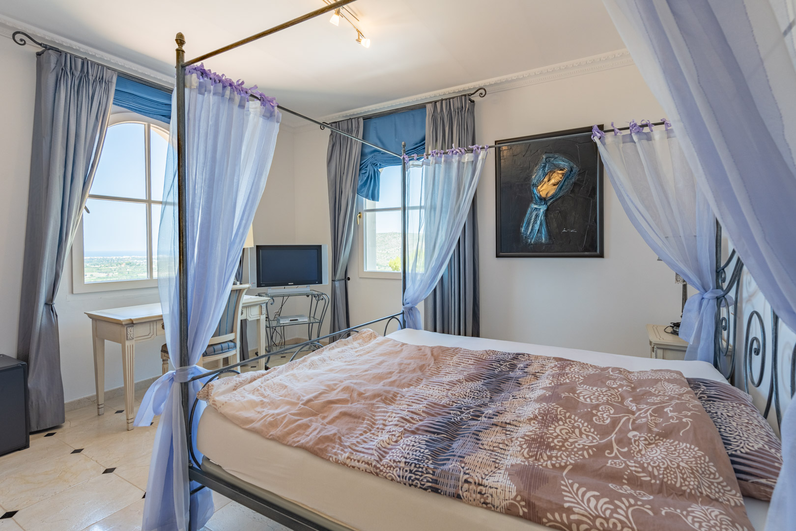Bedroom featuring a metal four-poster bed with sheer drapes, polished marble flooring with black accents, and a workspace overlooking the Mediterranean landscape.