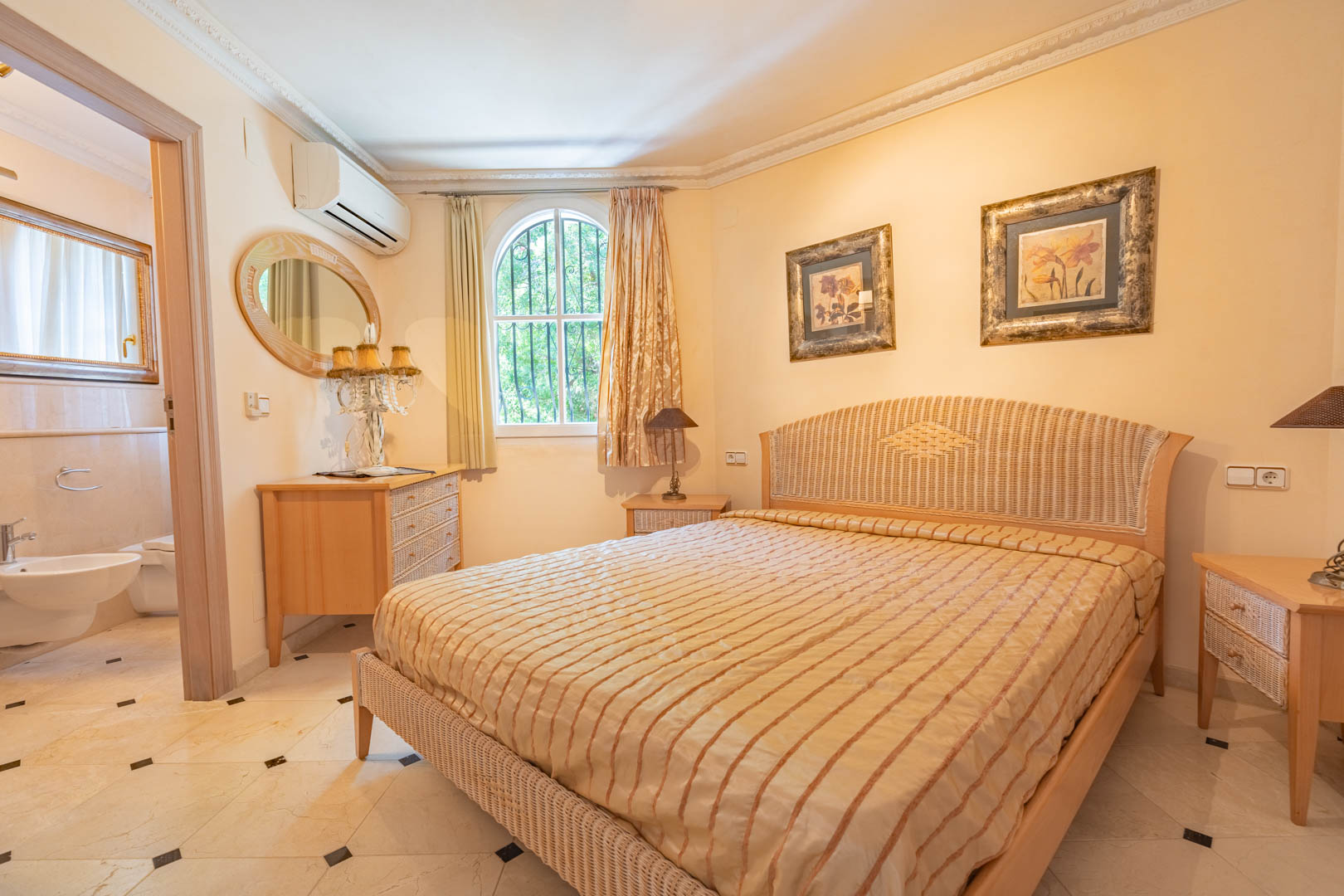Bedroom featuring a rattan bed frame, marble flooring with black accents, and an en-suite bathroom. Equipped with split air conditioning and a classic arched window.