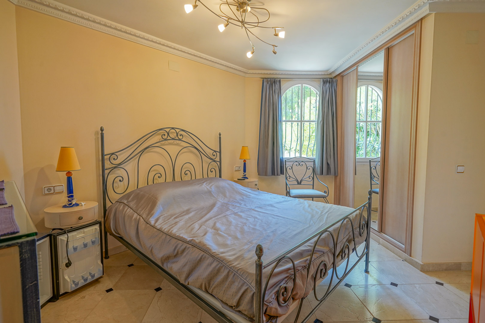 Bedroom featuring a wrought iron bed frame, marble tile flooring with black accents, built-in wardrobes, and an arched window with garden views.