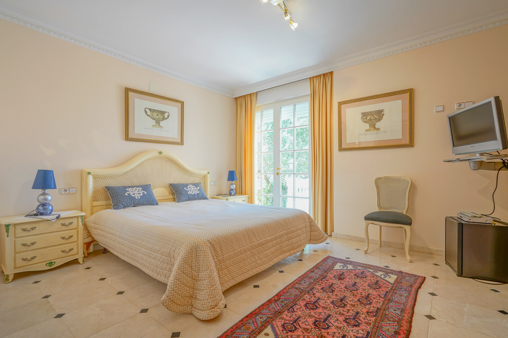 Bright bedroom featuring marble flooring with dark accents and classic cream furniture. Large glass doors provide natural light and direct access to the outdoor areas.