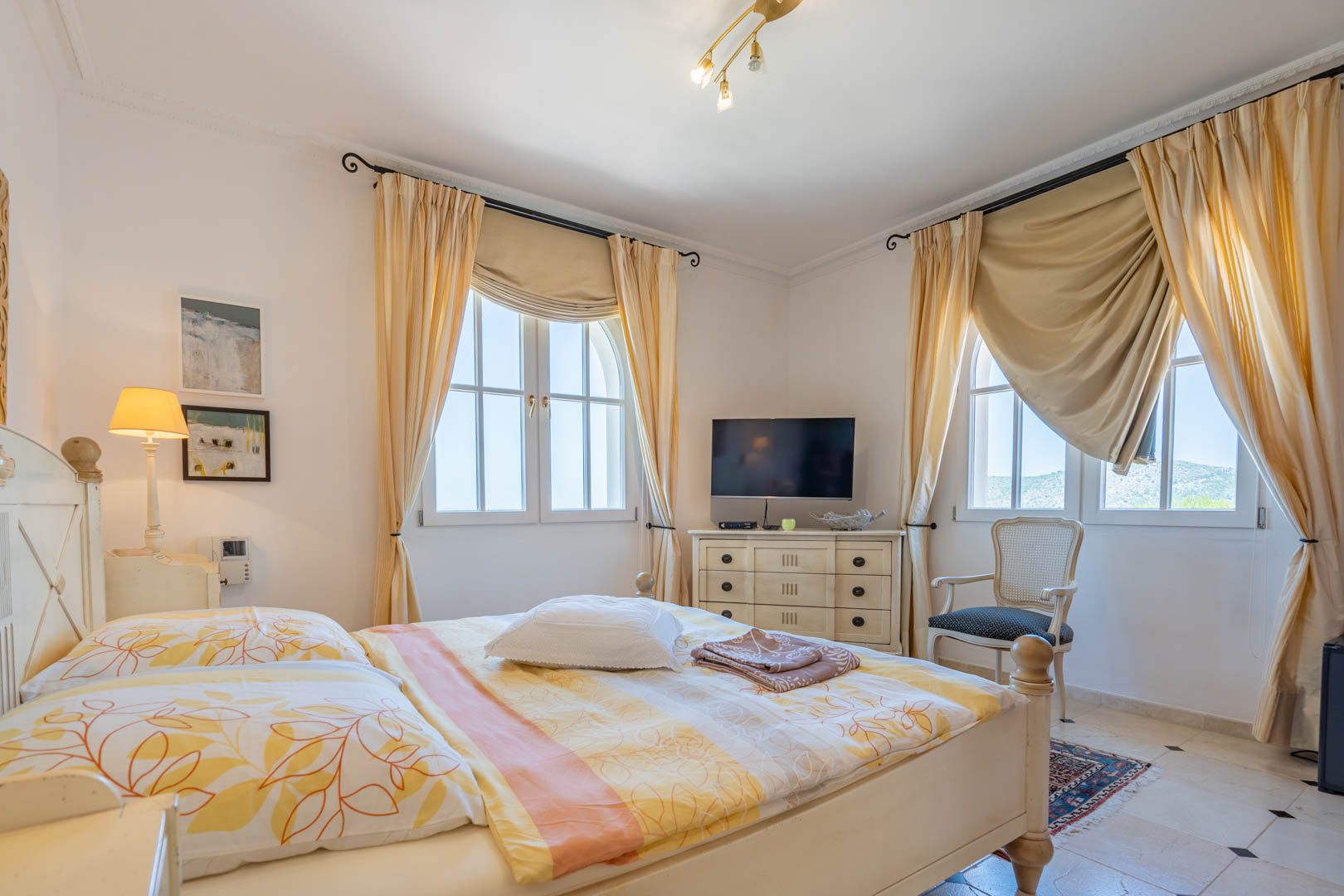 Bright bedroom featuring white wooden furniture, arched windows with cream drapery, and tiled flooring with dark accents. Includes a dedicated TV area and a classic armchair.