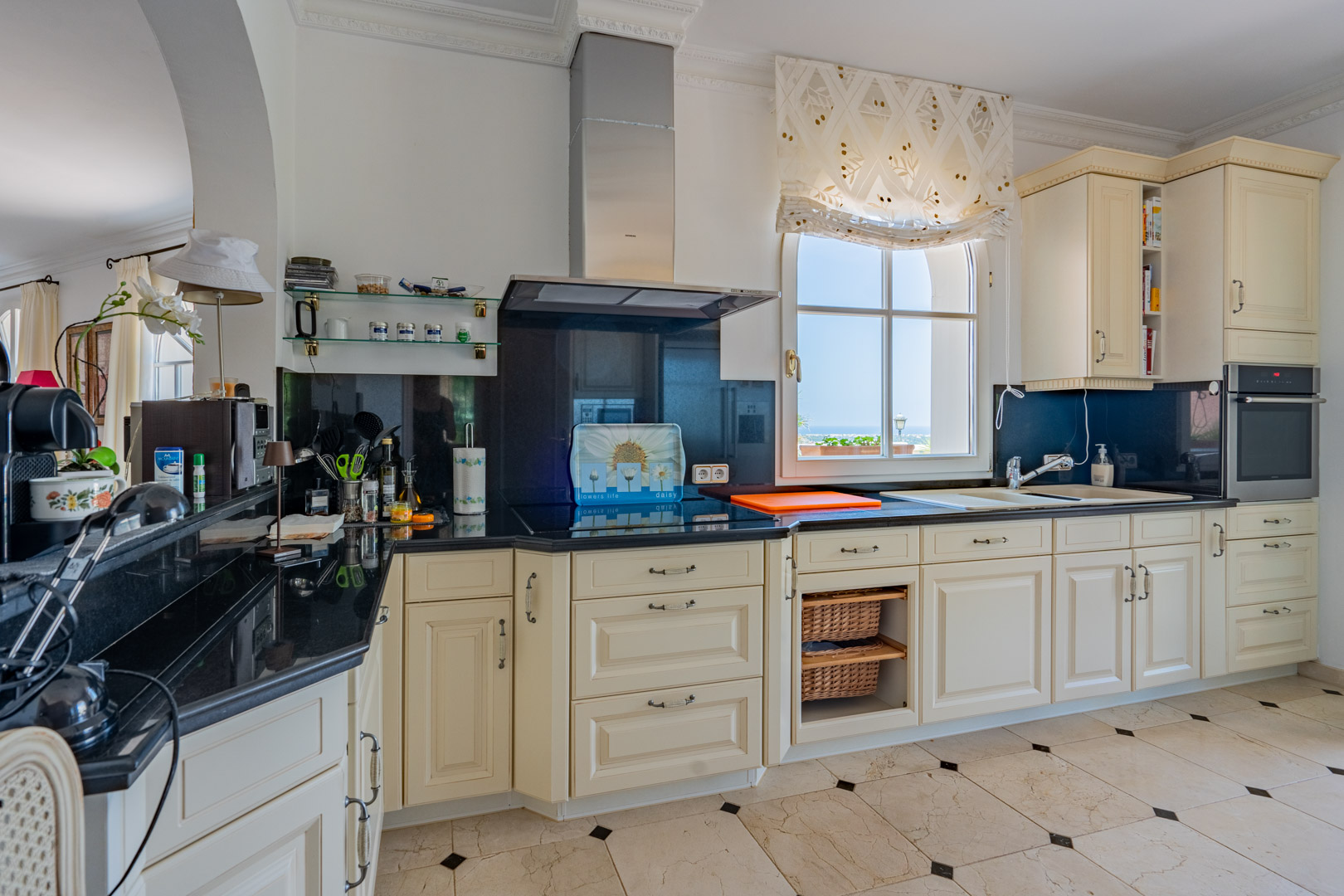 Classic kitchen featuring cream cabinetry, black granite countertops, and marble floors with dark accents. Includes integrated appliances and a window with coastal views.