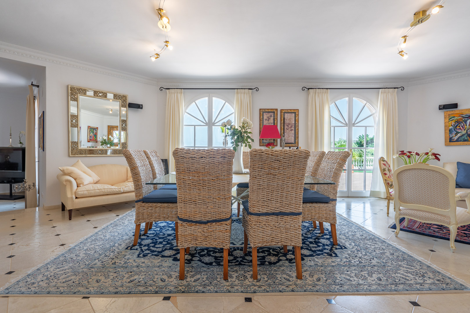 Elegant dining area featuring a glass table and wicker chairs on a patterned rug. Arched windows provide abundant natural light and direct access to the outdoor terrace.