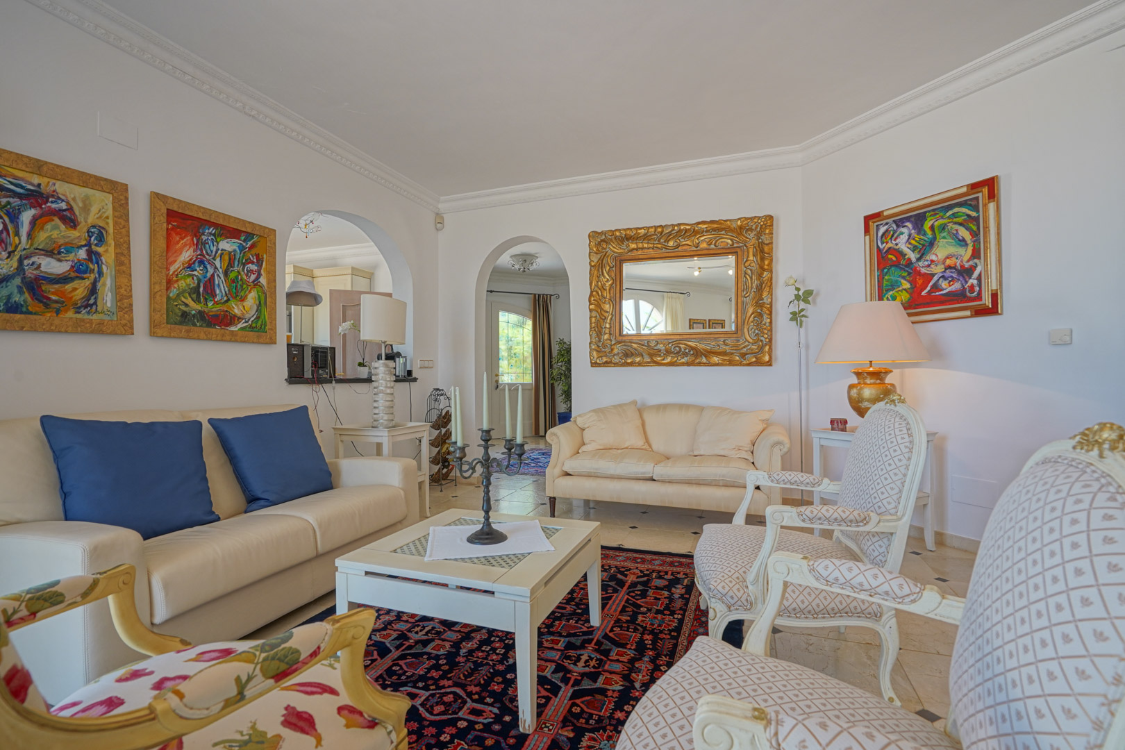Elegant living room featuring classic armchairs, leather sofas, and architectural arches. The space is accented by ornate gold-framed mirrors, vibrant artwork, and crown molding.