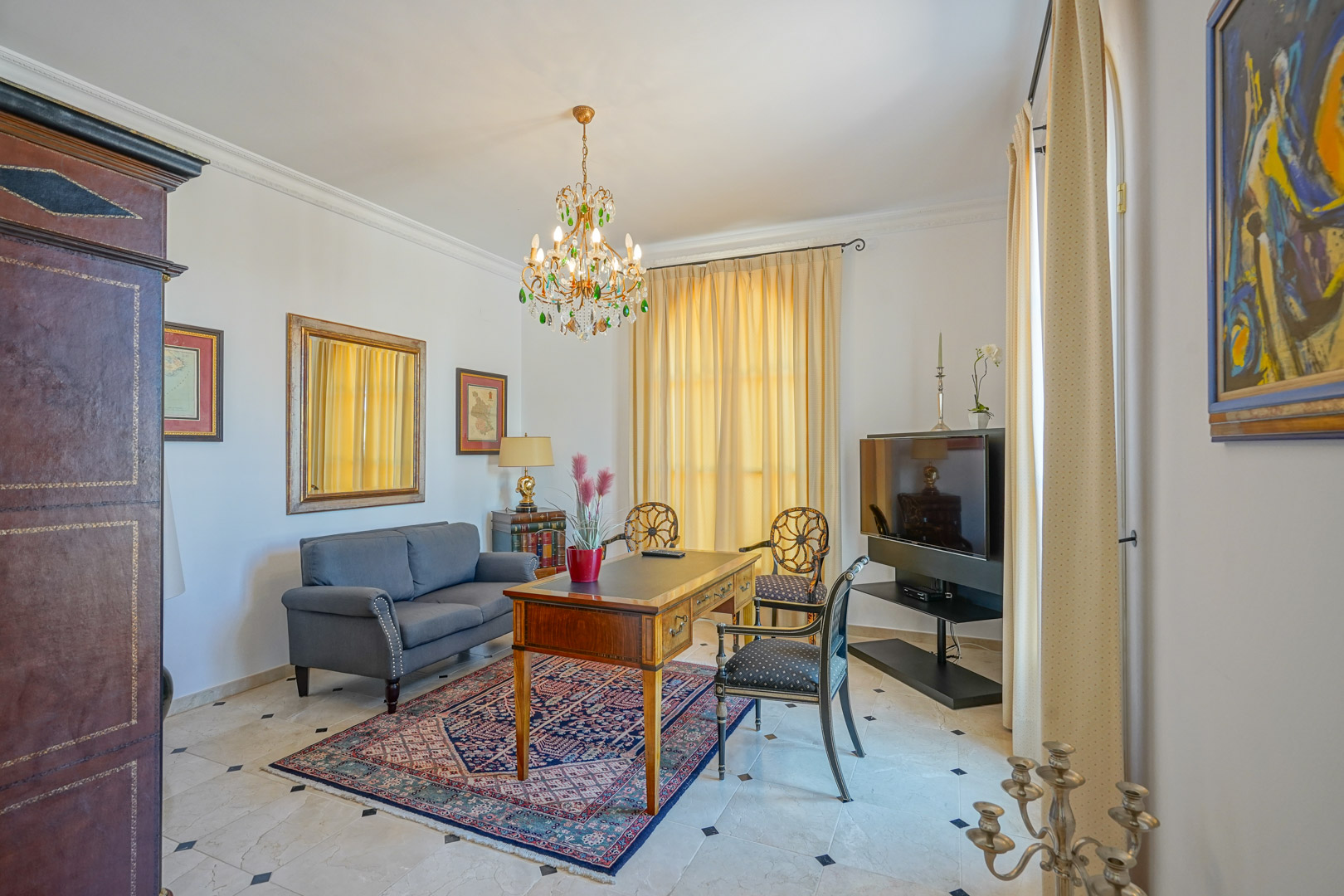 Elegant study featuring marble flooring with diamond inserts, a classic wooden desk, grey sofa, and a crystal chandelier with green accents.