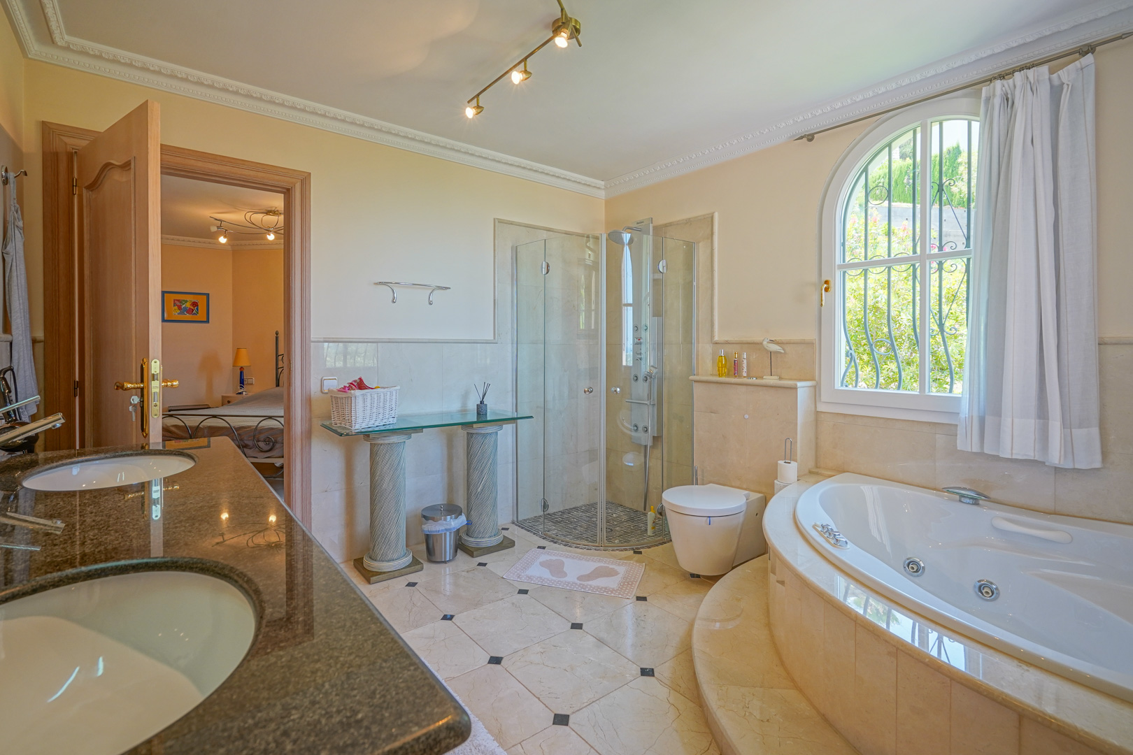 En-suite bathroom featuring a circular whirlpool tub, glass-enclosed shower, and double granite vanity. Includes marble flooring and an arched window overlooking the garden.