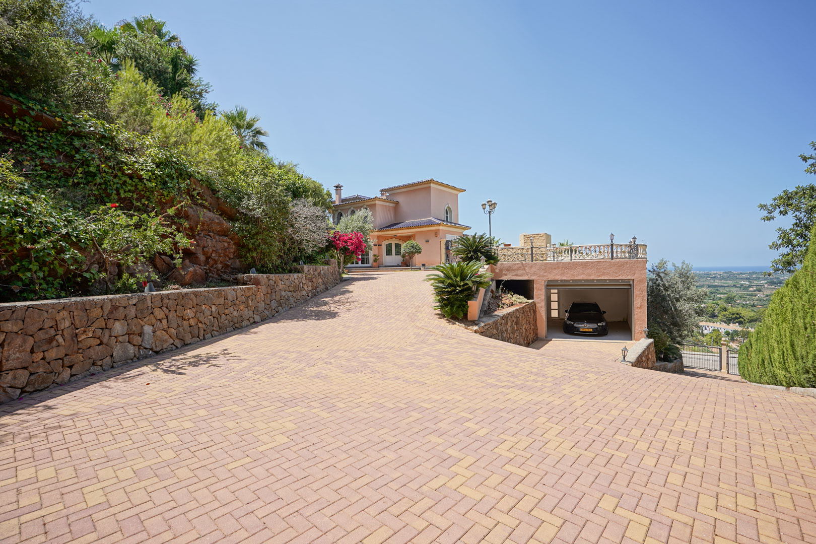 Extensive paved driveway leading to the villa and integrated garage, featuring natural stone walls, lush landscaping, and elevated views of the Mediterranean coastline.