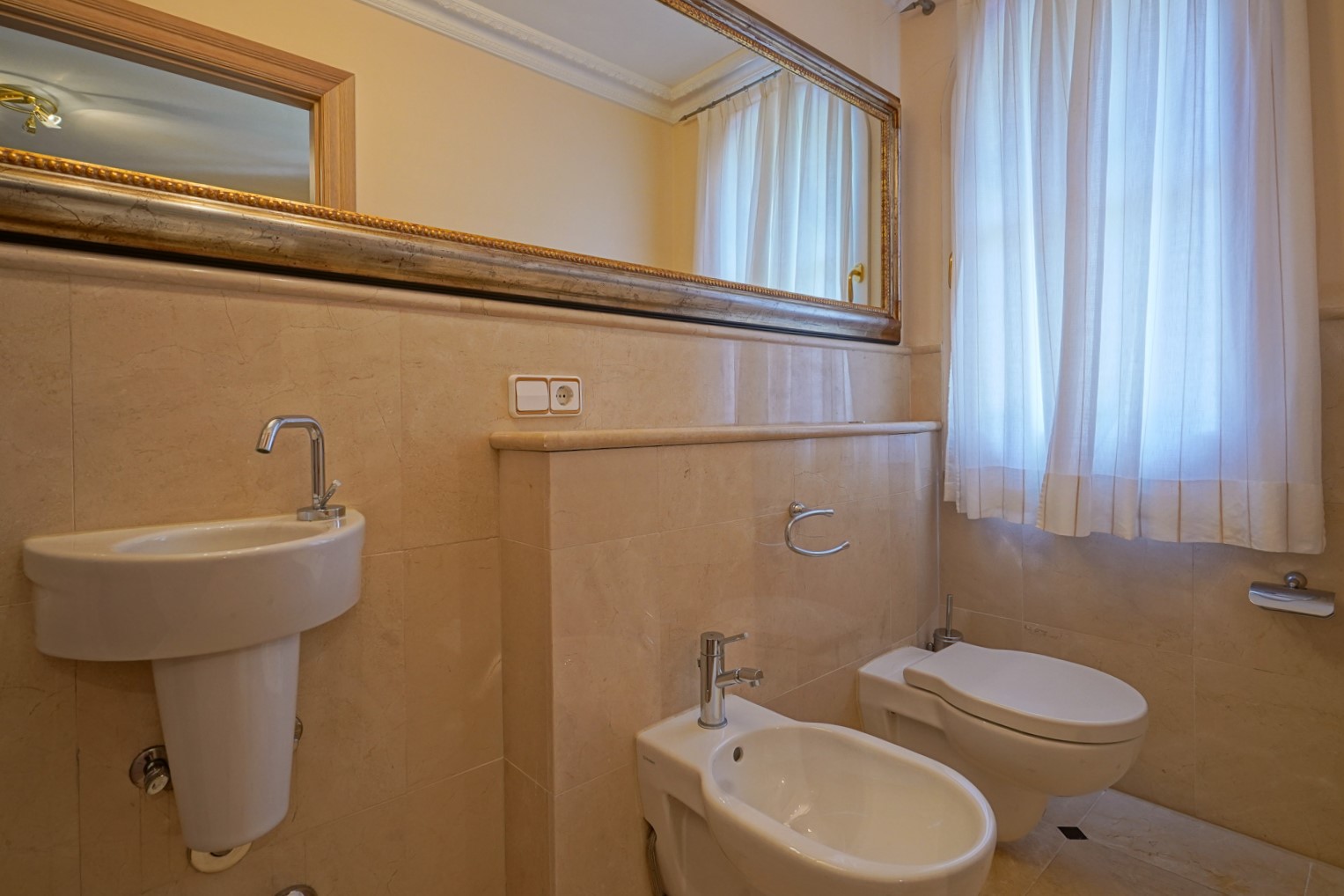 Guest bathroom featuring cream marble tiling, a pedestal sink, bidet, and toilet. A large framed mirror and window with soft curtains enhance the natural light and space.