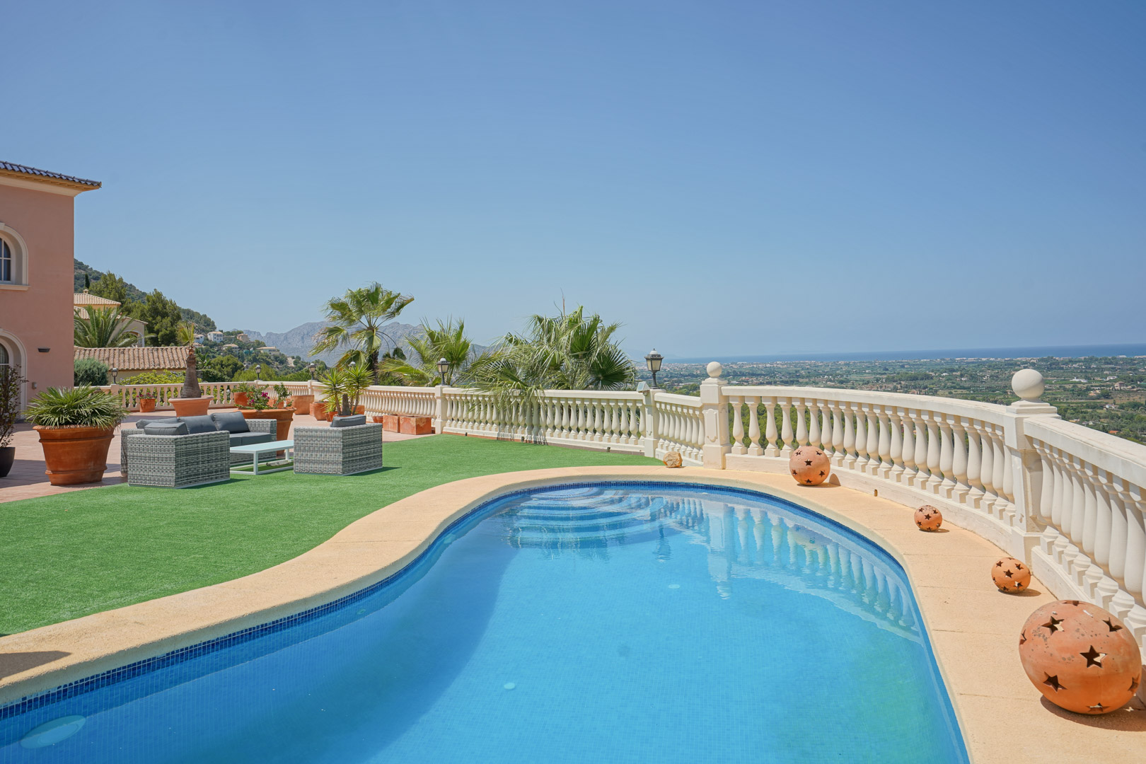 Kidney-shaped pool featuring panoramic sea and valley views, bordered by a classic stone balustrade, artificial turf, and a comfortable wicker lounge area.