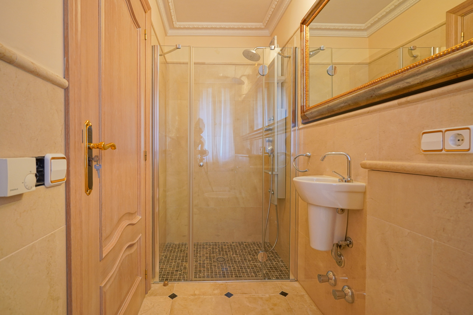 Marble-clad bathroom featuring a walk-in shower with mosaic flooring and glass enclosure. Equipped with a compact pedestal sink and an ornate gold-framed mirror.