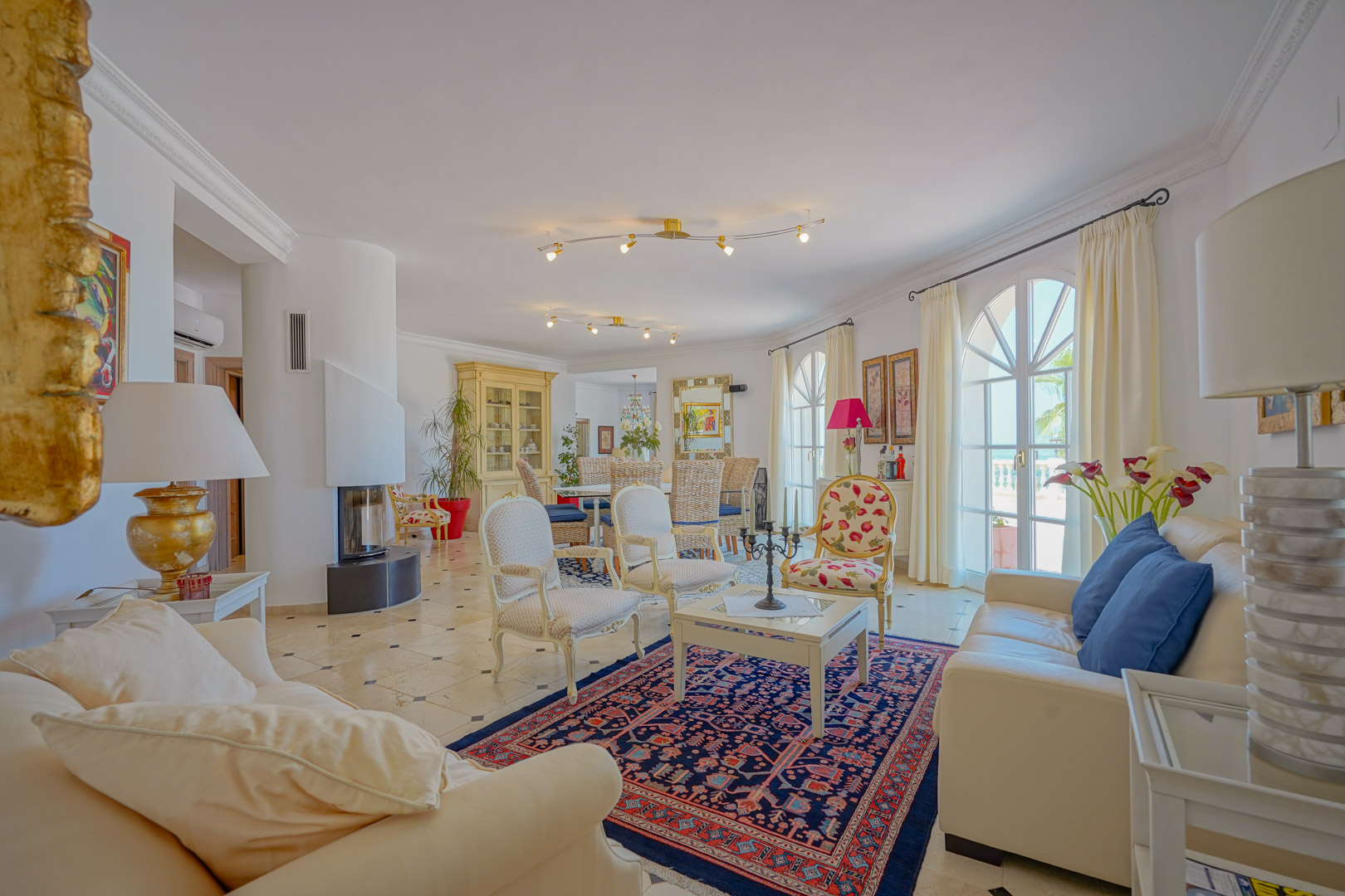 Open-plan living area featuring a modern curved fireplace, marble flooring with decorative inserts, and arched French doors opening to the terrace.