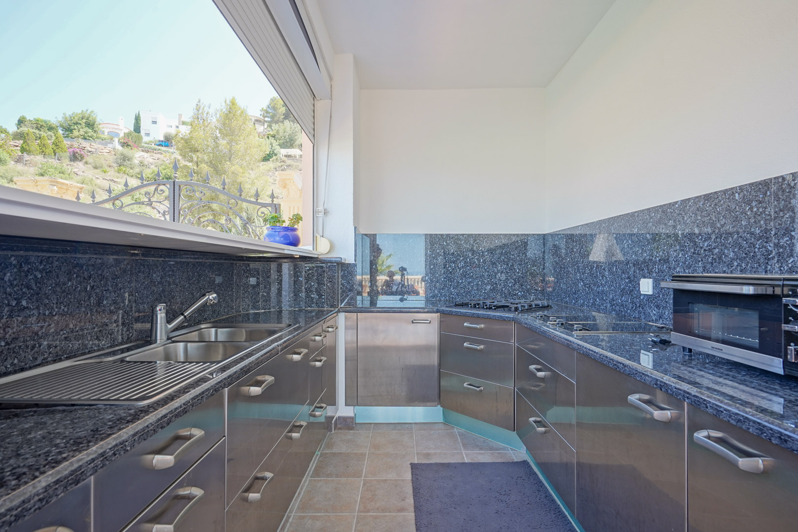 Outdoor kitchen featuring dark granite countertops, stainless steel cabinetry, and a double sink, positioned by a large window with views of the Mediterranean hillside.