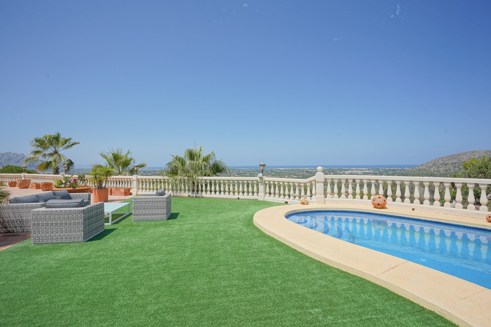 Outdoor terrace featuring a curved swimming pool, artificial lawn, and wicker lounge furniture overlooking the Mediterranean Sea and the La Sella valley.