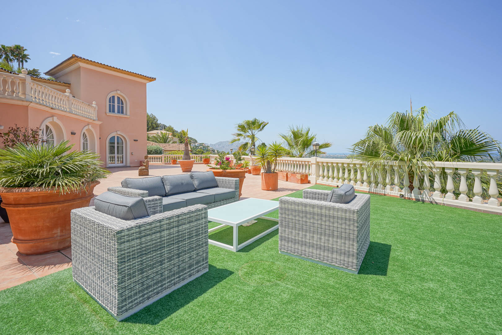 Spacious terrace featuring grey wicker lounge furniture on artificial turf, framed by terracotta planters and a classic balustrade with panoramic views.