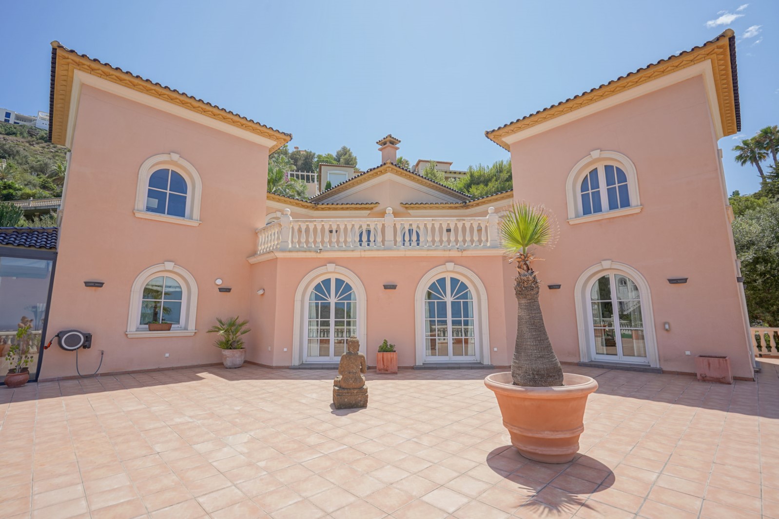 Symmetrical salmon-toned facade featuring arched windows, an upper terrace with white balustrades, and a spacious terracotta-tiled courtyard with a central stone statue.