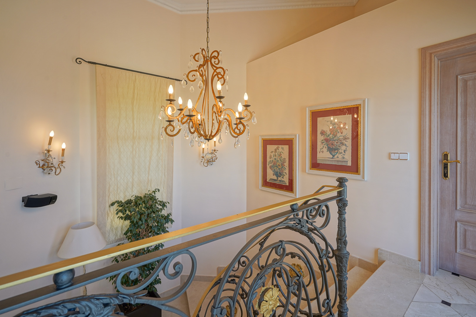 Upper landing featuring a wrought iron staircase railing with gold accents, a crystal chandelier, and Bose wall speakers against neutral-toned walls.