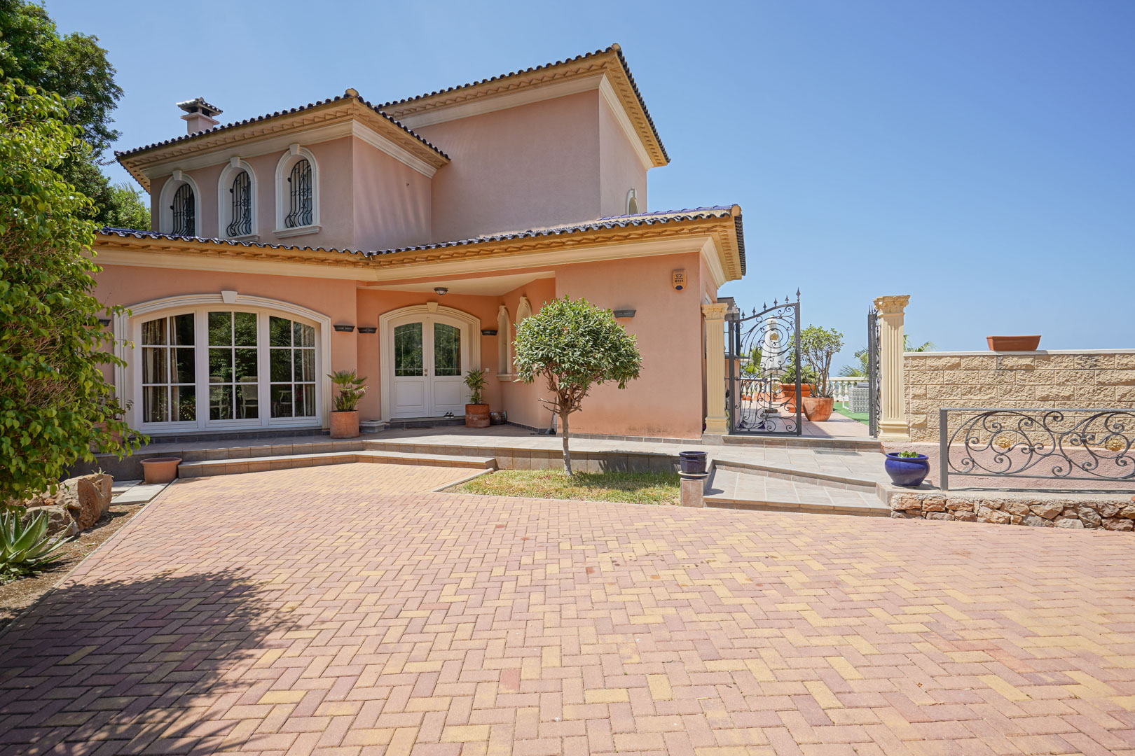 Villa exterior featuring a paved herringbone driveway, arched windows with wrought-iron details, and a white double entrance door under a clear blue sky.