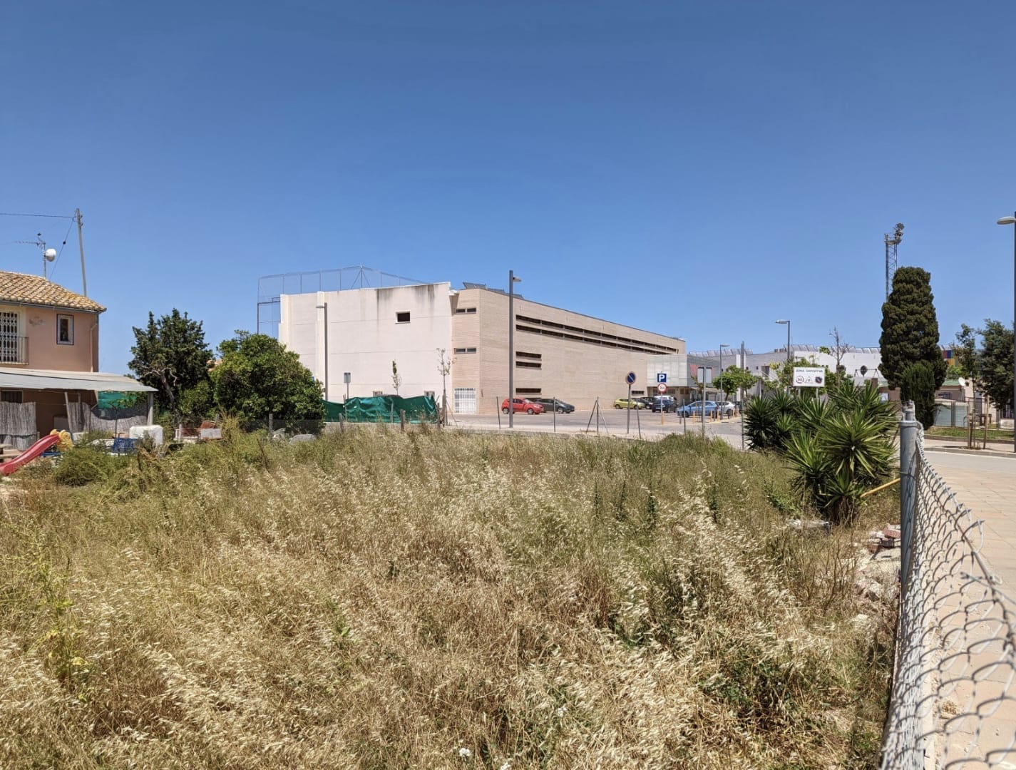 Flat urban plot with natural vegetation, located directly across from a modern sports facility and public parking under a clear sky.