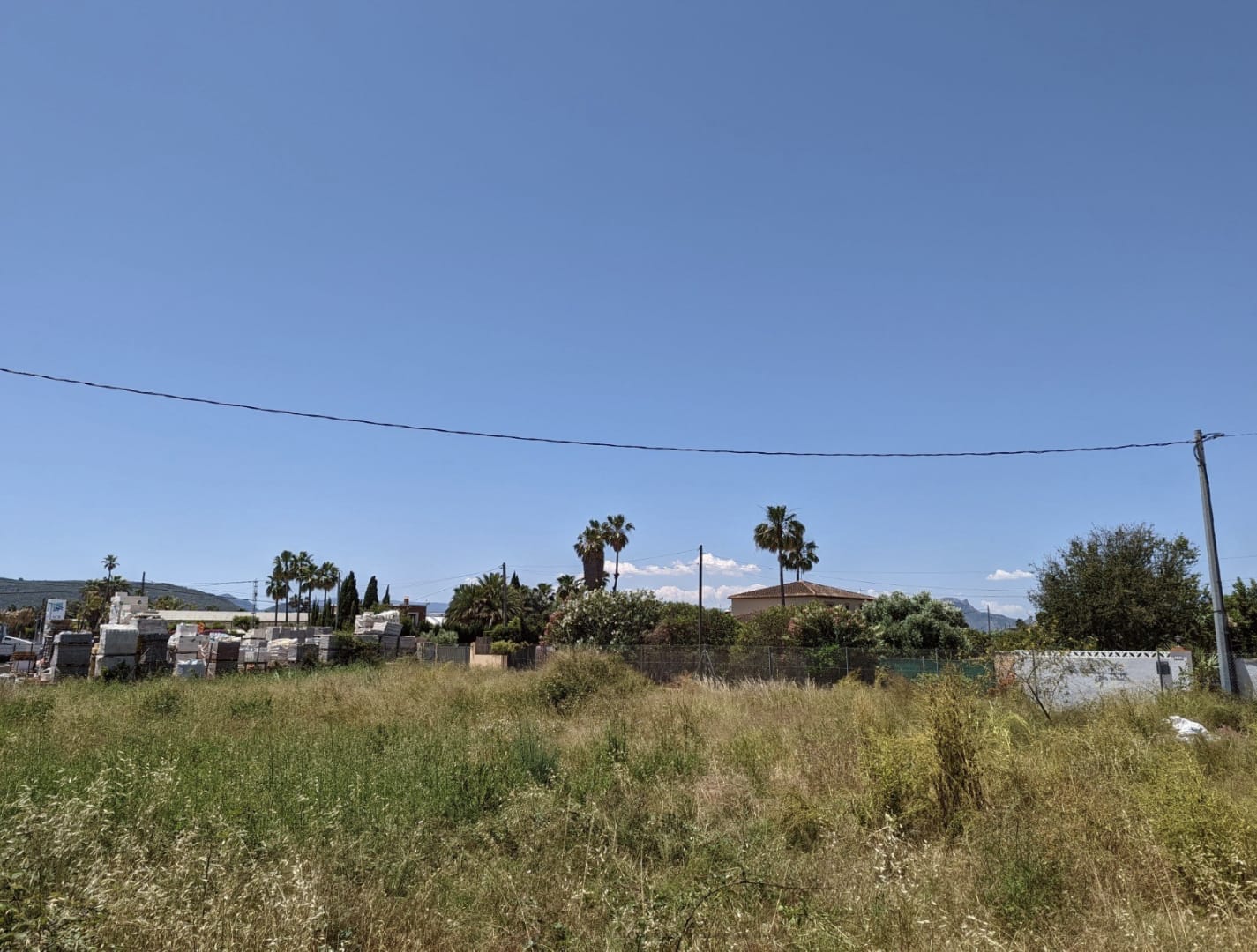 Level plot with wild grass and stored construction materials. Clear views of palm trees and nearby residential structures under a bright blue sky.