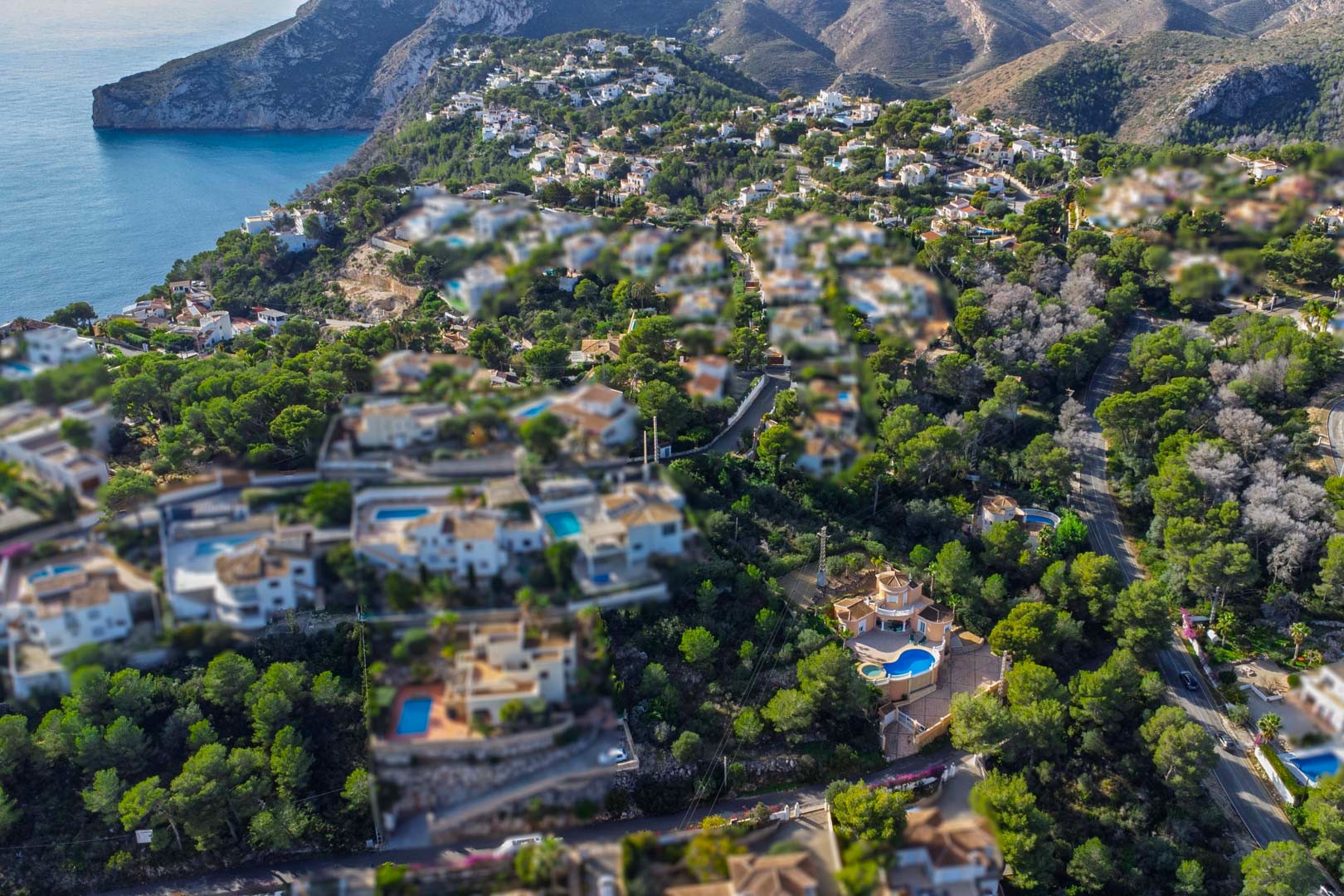 Aerial perspective of a hillside neighborhood in Javea, showcasing Mediterranean villas with private pools nestled among lush pine trees near the coastline.