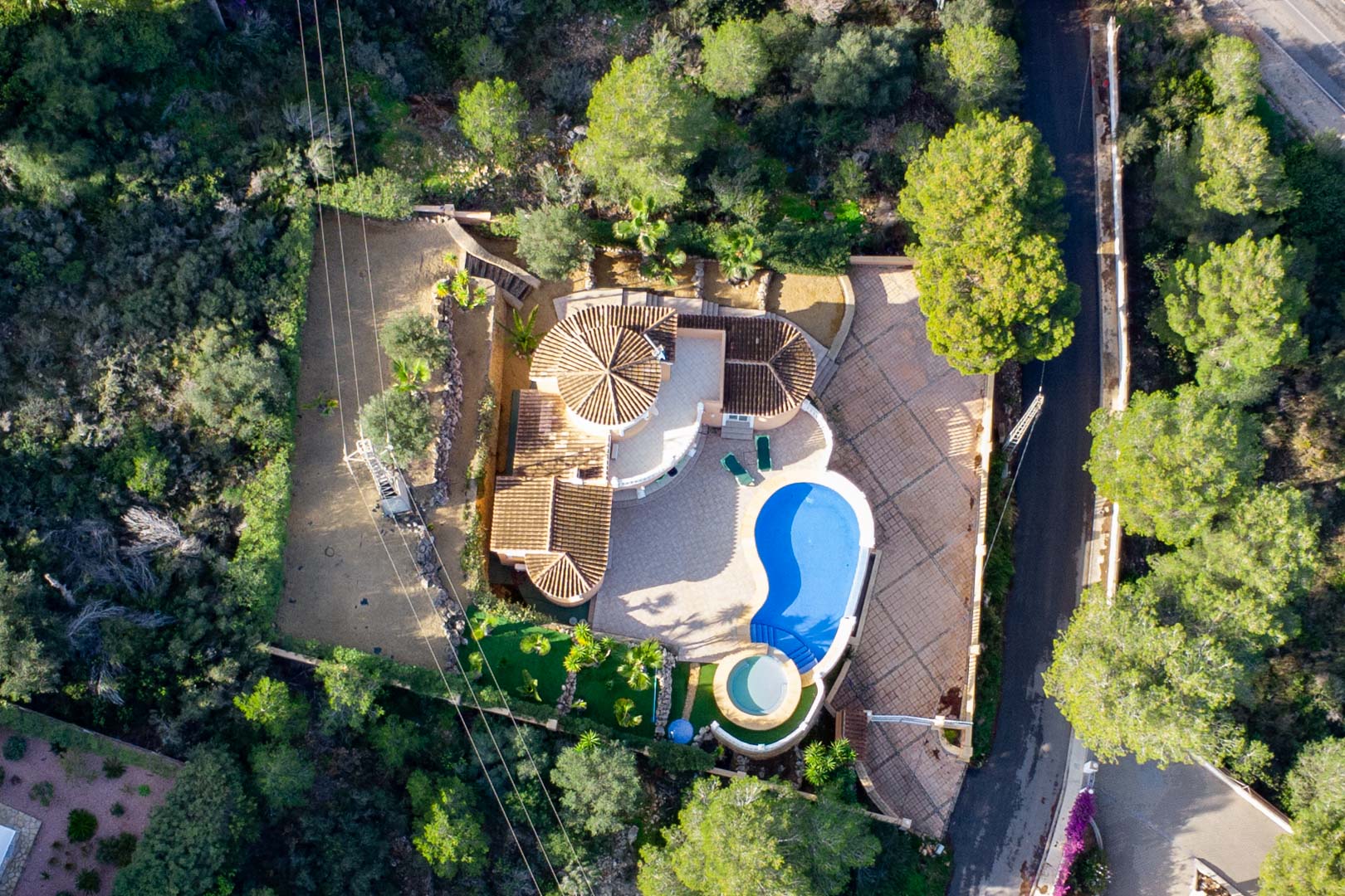 Aerial view of the villa showing the terracotta tiled roof, kidney-shaped swimming pool, and adjacent circular jacuzzi surrounded by a dense Mediterranean forest.