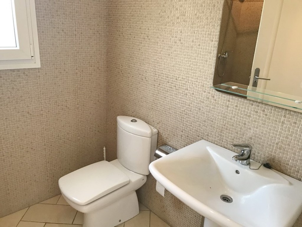 Bathroom featuring beige mosaic wall tiles, a white ceramic pedestal sink, and a matching toilet. Includes a mirror with a glass shelf and neutral-toned floor tiling.