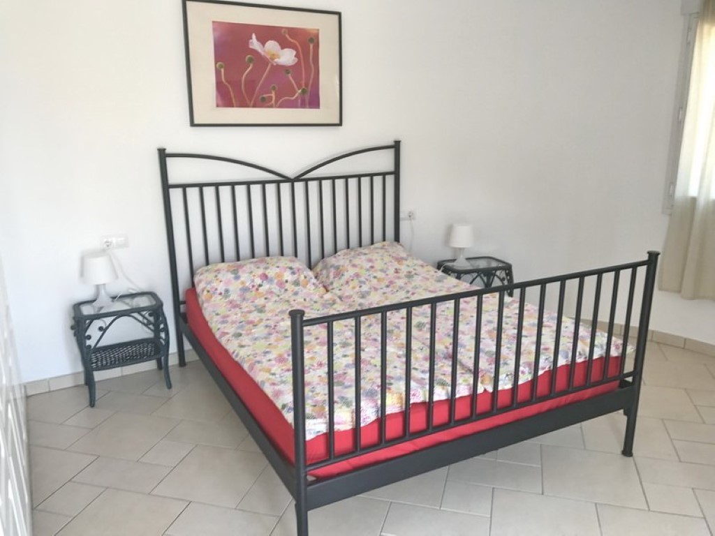 Bedroom featuring a black wrought-iron bed frame, matching wicker nightstands, and light tiled flooring. Natural light complements the white walls and floral decor.