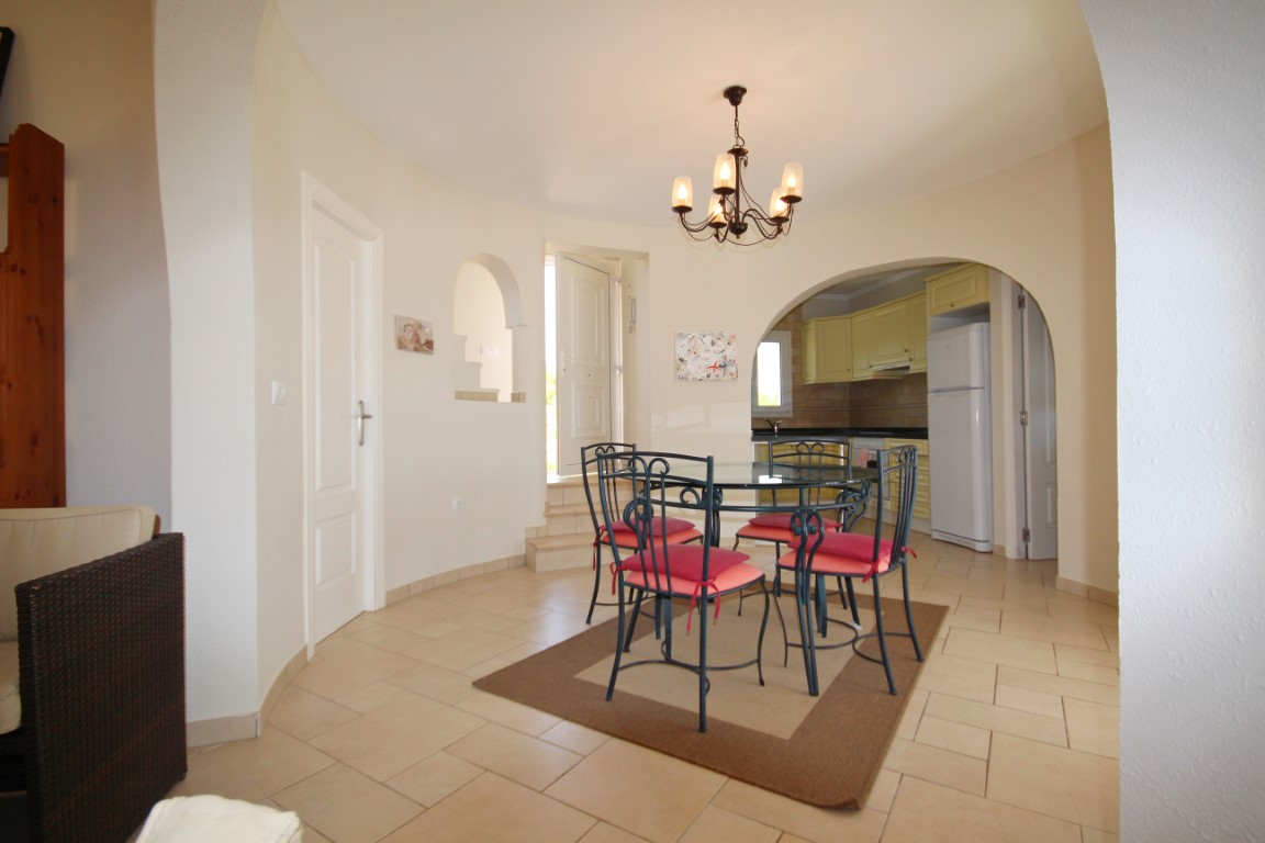 Bright dining area featuring a wrought iron table with glass top, arched transitions to the kitchen, and neutral tiled flooring under a classic chandelier.