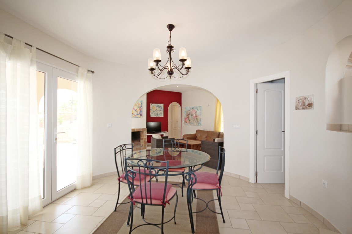 Dining space featuring a circular glass table and wrought iron chairs, with a classic chandelier and an archway leading to a living room with a red accent wall.