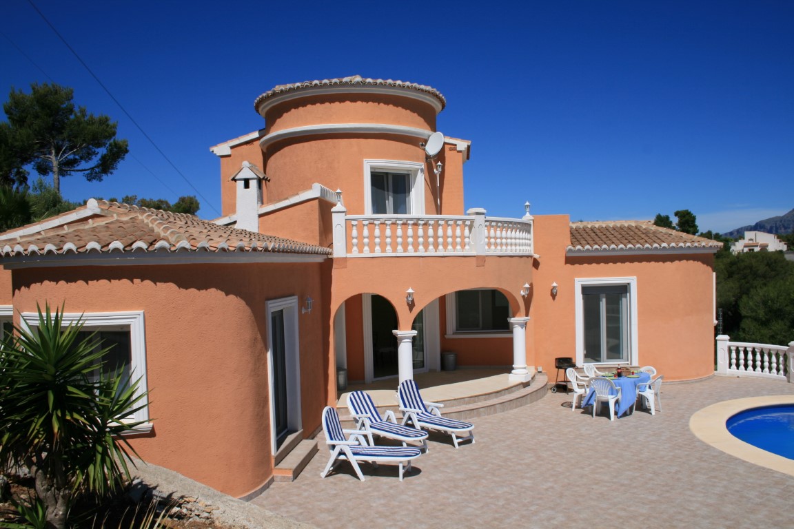 Exterior of a terracotta villa featuring a rounded tower, arched porch, and a tiled terrace with sun loungers and a private pool area.