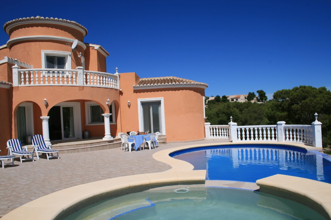 Exterior view of a terracotta villa featuring a kidney-shaped pool and integrated jacuzzi, surrounded by a tiled terrace with white balustrades and arched porches.