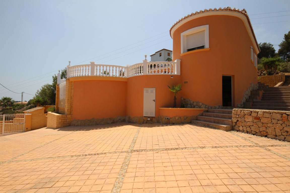 Exterior view of the villa featuring a terracotta facade, curved architecture, and a spacious tiled driveway. Stone retaining walls and white balustrades define the terrace levels.