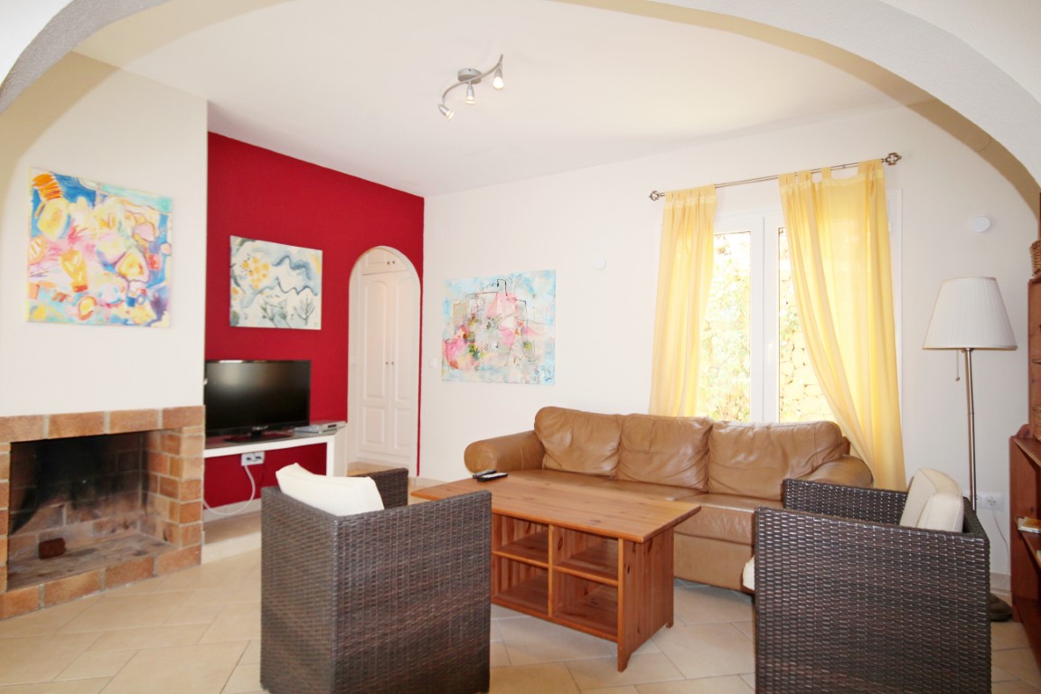 Living room featuring a brick fireplace, red accent wall, and tan leather sofa. Includes wicker armchairs, a wooden coffee table, and natural light through yellow curtains.