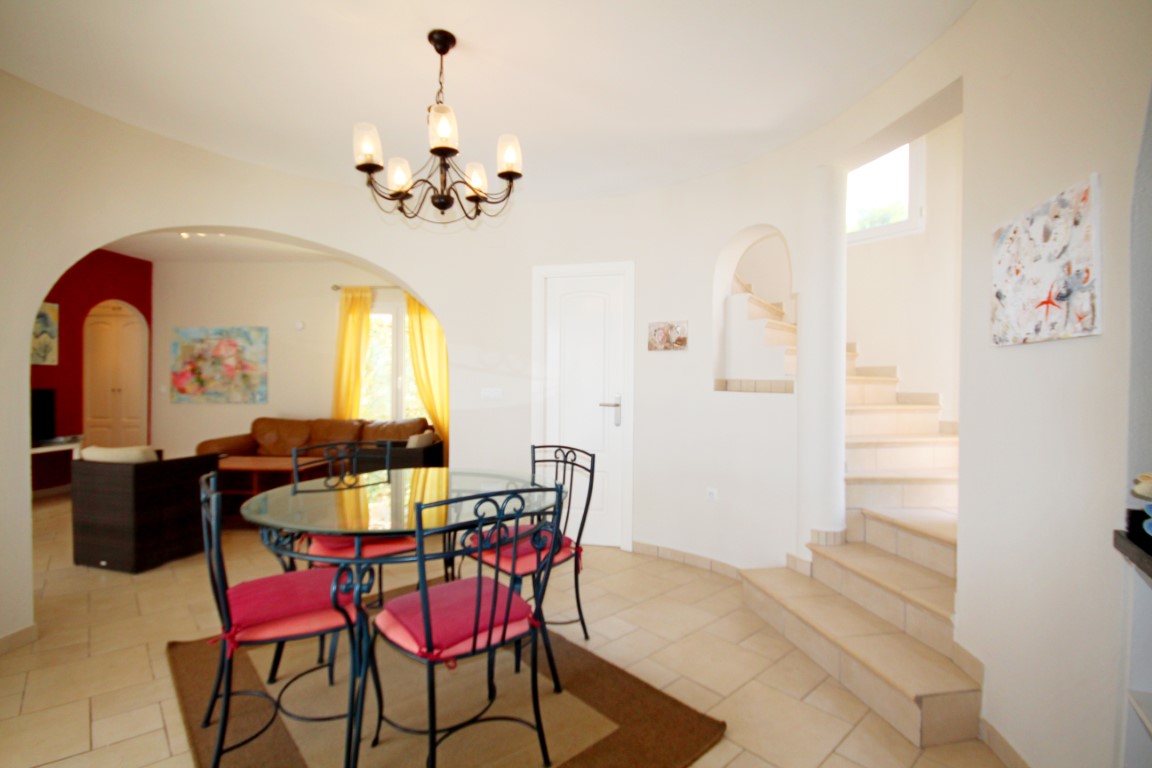 Open-plan dining area featuring a glass-top table with wrought iron chairs, connected to the living room via a wide archway and adjacent to the tiled staircase.