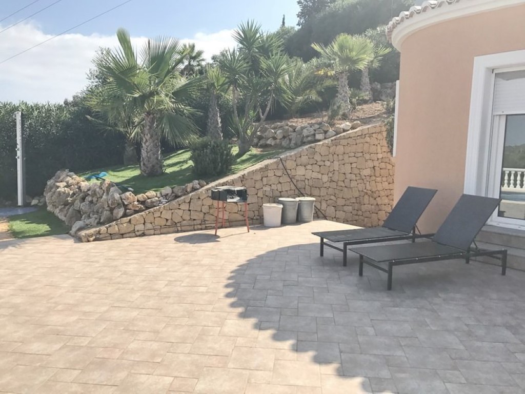 Paved outdoor terrace featuring black sun loungers, a natural stone retaining wall, and a landscaped Mediterranean garden with palm trees.