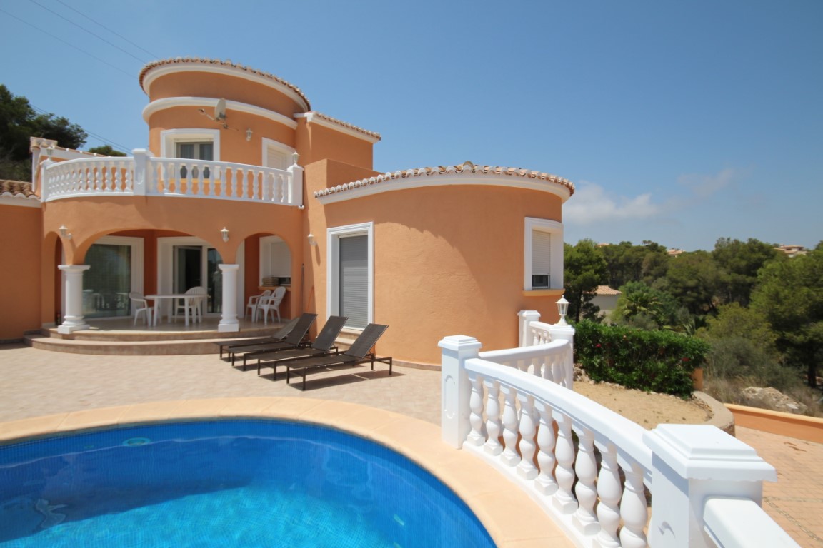 Villa exterior featuring a private blue-tiled pool, sun loungers, and a terracotta facade. The architecture includes white balustrades and a shaded arched ground-floor terrace.