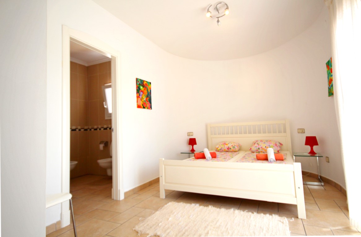 Bedroom featuring a curved wall, white wooden bed frame, and an integrated en-suite bathroom with neutral tiling.
