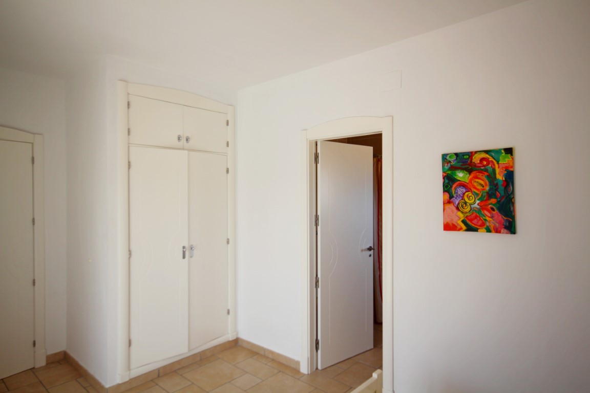Bright bedroom area featuring a white built-in wardrobe, rustic terracotta-style floor tiles, and a doorway leading to an adjacent room.